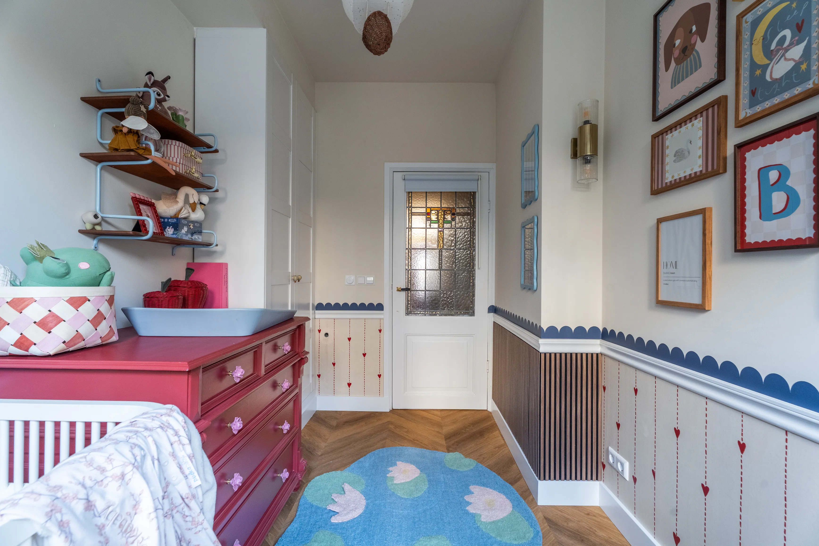 Een vloerkleed met waterlelies en een bessenrode commode onder een geschulpte blauwe rand en glas-in-lood deur