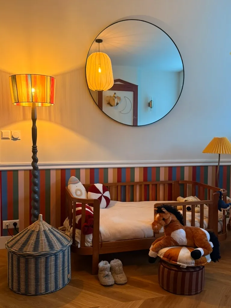 Een kinderbed onder een ronde spiegel, verlicht door een gestreepte vloerlamp