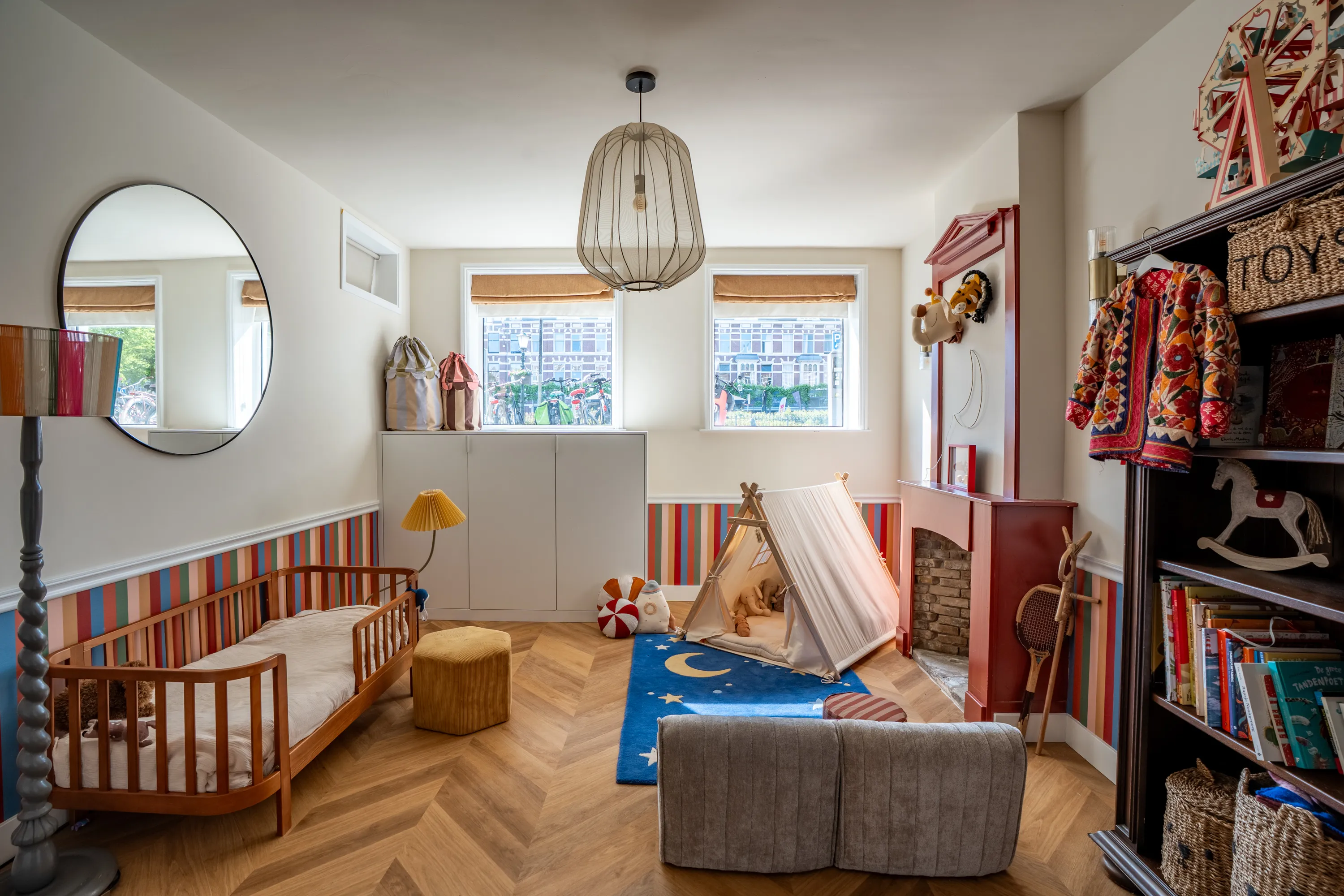 Een kinderbed onder een ronde spiegel, verlicht door een gestreepte vloerlamp